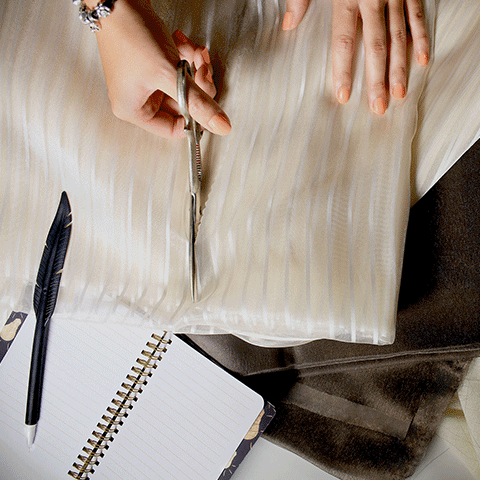 Two hands, one hand with a pair of scissors cutting a sheer fabric and a notebook with a feather pen	